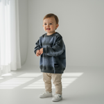 Child wearing a gray tie-dye sweatshirt and beige pants standing in a bright room.